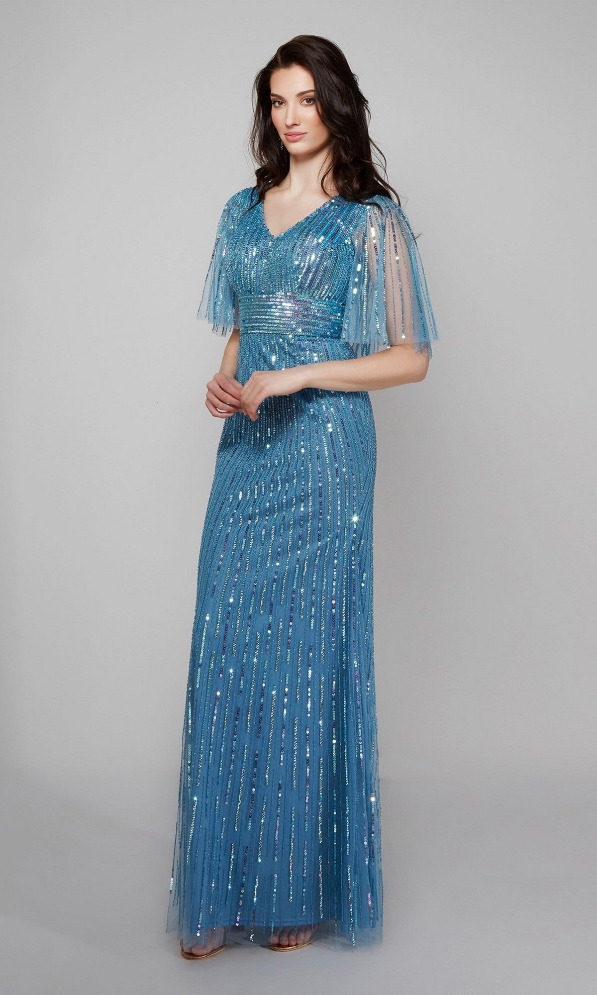 A woman wears the Alyce 27599 long formal dress, featuring blue beaded sequins and short sheer sleeves, standing against a plain light gray background.